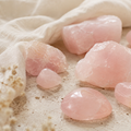 Polerade och råa rosenkvartsstenar i mjuka rosa toner på ljus sten och beige linnetyg.